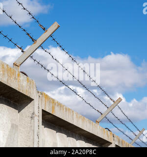 Stacheldraht in drei Linien oben auf einem alten Beton Zaun auf graues Metall Beiträge mit blauem Himmel und weißen Wolken Hintergrund Rost gestreckt Stockfoto