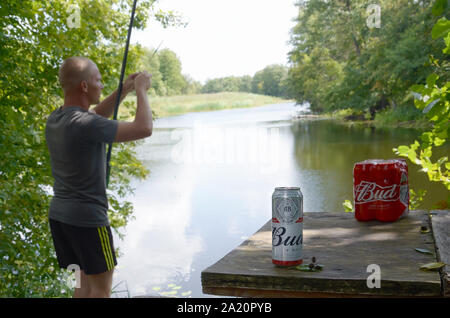 Kharkov, Ukraine - August 10, 2019: Budweiser Bud Bierdosen pack auf alten Tisch und Fischer am Fluss für den Hintergrund. Budweiser ist einer der beliebtesten Stockfoto