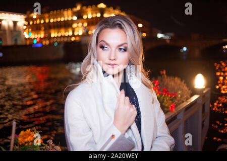 Stilvolle und elegante junge Frau in einem beigen Mantel auf der Stadt am Wasser. Nacht Stadt im Herbst. Portrait von charmante Blondine in der Nacht im Freien. Stockfoto