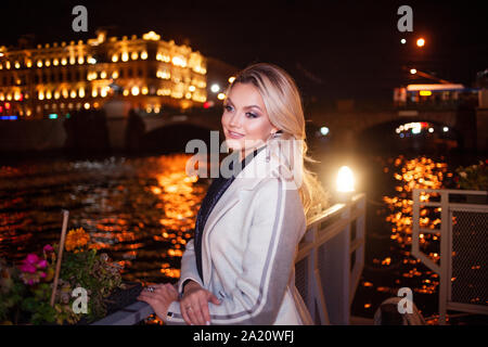 Stilvolle und elegante junge Frau in einem beigen Mantel auf der Stadt am Wasser. Nacht Stadt im Herbst. Portrait von charmante Blondine in der Nacht im Freien. Stockfoto