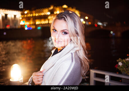 Stilvolle und elegante junge Frau in einem beigen Mantel auf der Stadt am Wasser. Nacht Stadt im Herbst. Portrait von charmante Blondine in der Nacht im Freien. Stockfoto