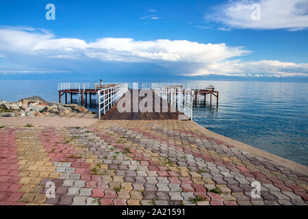 Pier am Issyk-Kul See auf dem Hintergrund einer Bergkette mit schneebedeckten Gipfeln und bewölktem Himmel. Kirgisistan Stockfoto