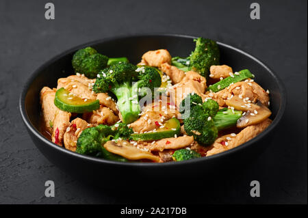 Hunan Chicken in schwarz Schüssel am dunklen Schiefer Hintergrund. Hunan Chicken ist Chinesisch oder indisch-chinesische Küche takeaway Gericht mit Brokkoli, Zucchini, Shiitake Stockfoto