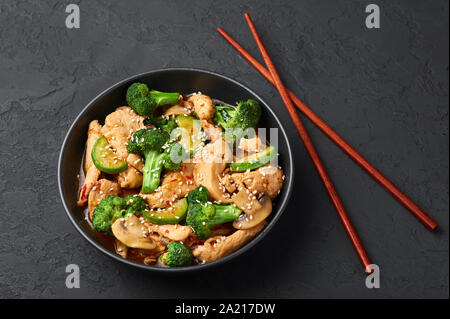 Hunan Chicken in schwarz Schüssel am dunklen Schiefer Hintergrund. Hunan Chicken ist Chinesisch oder indisch-chinesische Küche takeaway Gericht mit Brokkoli, Zucchini, Shiitake Stockfoto