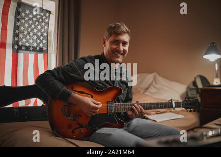 Porträt eines lächelnden jungen männlichen Musiker Gitarre spielen in seinem Schlafzimmer Blick von der Kamera Stockfoto