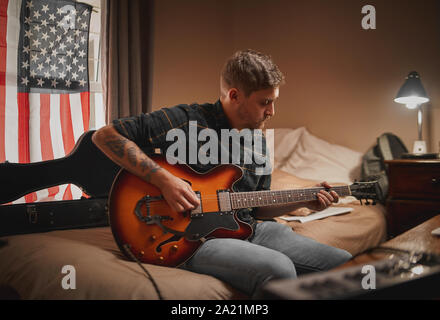 Konzentrierter junger Mann in Freizeitkleidung sitzt auf dem Bett Gitarre zu spielen, abends Stockfoto