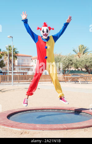 Ein furchtsames Clown trägt einen bunten gelben, roten und blauen Kostüm Springen auf einem Trampolin in der freien öffentlichen Spielplatz Stockfoto