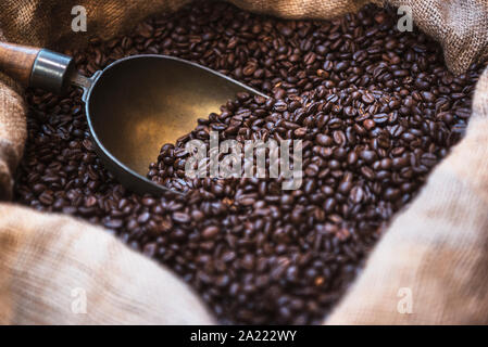 In der Nähe von frisch gerösteten Arabica Kaffeebohnen mit einer alten Schaufel in einem Jute Tasche. Kaffeebohnen in offenen Sack. Geröstete Kaffeebohnen schwarzen Hintergrund. Stockfoto