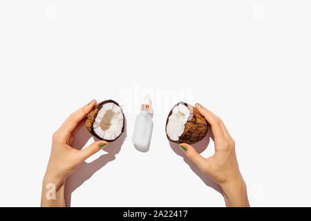 Bio Kokosöl Flasche zwischen Woman's Hände, die zwei Hälften eines frisch gerissenen Coconut auf weißem Hintergrund, flach. Stockfoto