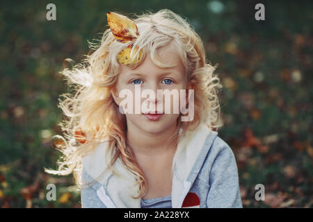 Schöne hübsche Vorschule Kaukasischen blonde Mädchen mit Blätter im Herbst in langen Haaren sitzen auf Gras in die Kamera schauen. Portrait von Lovely cute adorable Stockfoto