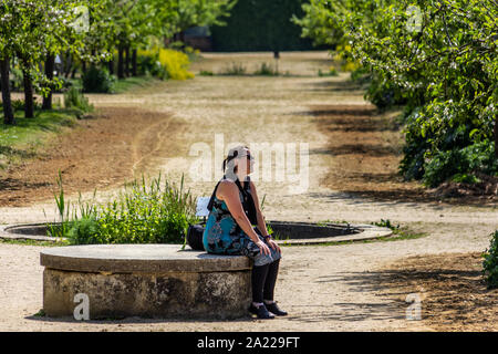 Eine Frau sitzt nachdenklich auf einer konkreten Bank in einem Hain von Bäumen im Garten in der Nähe von einem Brunnen Stockfoto