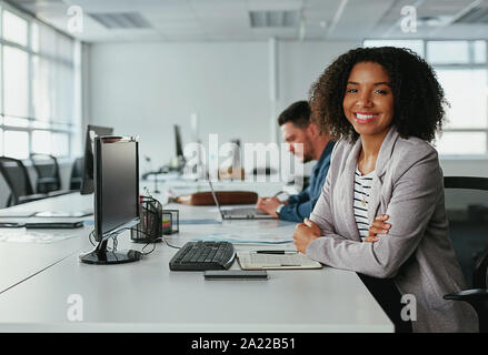 Erfolgreiche lächelnden jungen Geschäftsfrau mit verschränkten Armen auf Kamera und männlicher Kollege arbeitet an einem Schreibtisch im Hintergrund Stockfoto