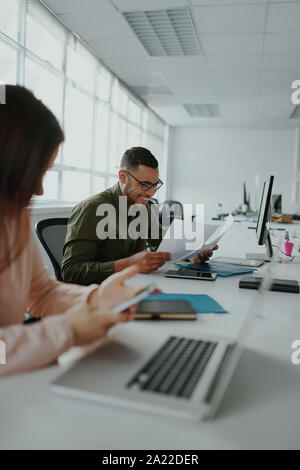 Erfolgreiche junge Unternehmer auf der Suche nach Dokumenten, während seine Kollegin mit Smartphone mit Laptop auf dem Schreibtisch - echte Menschen Stockfoto
