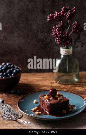 Hausgemachte vegan Chocolate Brownie, frisch gebackene, mit Schokoladensauce und eine Kirsche, auf blauen Platte und mit Heidelbeeren serviert gekrönt. Holztisch, da Stockfoto