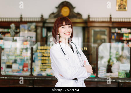 Selbstbewussten jungen Frau Apotheker mit einem reizenden Lächeln stand mit verschränkten Armen in der Apotheke Stockfoto