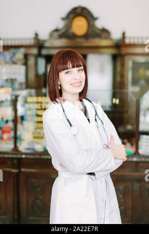 Selbstbewussten jungen Frau Apotheker mit einem reizenden Lächeln stand mit verschränkten Armen in der Apotheke Stockfoto