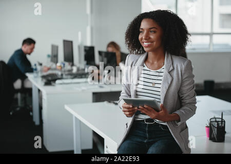 Happy nachdenkliche junge geschäftsfrau mit digitalen Tablet in der Hand lächelnd und Wegsehen vor Kollegen im Hintergrund Stockfoto