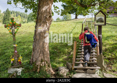Ein Wanderer klettert Holzstufen in Biala Woda in der Tatra, vorbei an einem Blech Jesus Kruzifix (eine populäre lokale Kopie von einem historischen Rutherian Kirche) in der Nähe von Jaworki, am 20. September 2019, in der Nähe von Szczawnica, Malopolska, Polen. Das Dorf eines florierenden Rutherian Gemeinschaft war einmal hier in Biala Woda wo über 100 Farmen wurden - die Reste davon sind noch heute zu sehen. Ein hölzernes Kreuz mit Christus Schneiden von Blech überlebte der Kultur. Ähnlich wie Kreuze und Kapellen kann in der Umgangssprache der Weißen Wasser in der Rumänisch, Ukrainisch oder Ost Slowakei gefunden werden. Stockfoto
