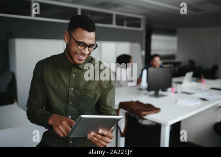 Fröhlicher junger Geschäftsmann mit digitalen Tablet lächelnd, während seine Kollegen im Hintergrund arbeiten Stockfoto