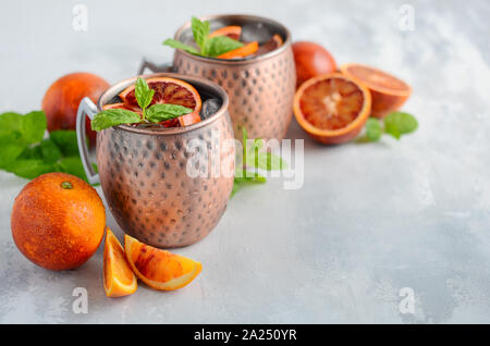 Blutorange Moscow Mule Alkohol Cocktail mit frischen Minzeblättchen und Eis in Kupfer Tassen auf grauem Beton Hintergrund. Stockfoto