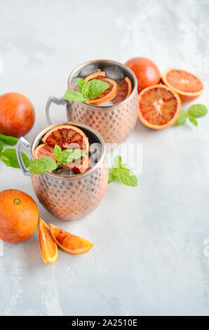 Blutorange Moscow Mule Alkohol Cocktail mit frischen Minzeblättchen und Eis in Kupfer Tassen auf grauem Beton Hintergrund. Stockfoto