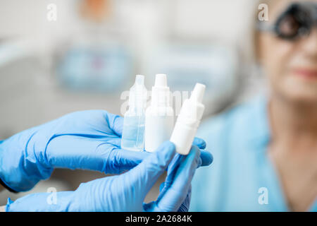 Krankenschwester in medizinische Handschuhe Holding weißen Flaschen oder Pipetten mit Auge Medizin mit älterer Patienten auf dem Hintergrund, Nahaufnahme, Flaschen mit leeren s Stockfoto