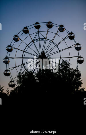 Silhouette einer Riesenrad bei Sonnenaufgang (Sonnenuntergang) der Sonne auf dem Hintergrund der Bäume und Gras. Schönen Hintergrund mit kopieren. Stockfoto