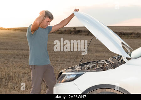 Ein Mann in der Mitte des Feldes Kontrolle der Motor seines aufgeschlüsselt Auto bei Sonnenuntergang. Lifestyle Konzept Stockfoto