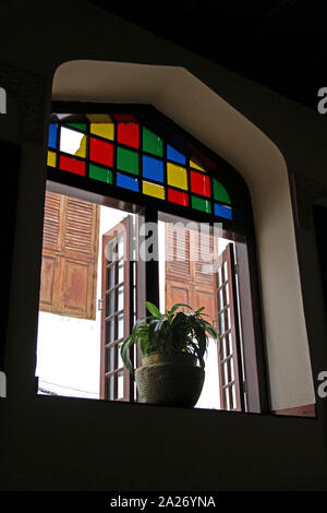 Ein Riss in einem Quadrat einer Glasmalerei doppeltes Fenster, mit einem potplant auf dem Schweller innerhalb einer Wohnanlage, Stone Town, Sansibar, Unguja Insel, Tansania. Stockfoto