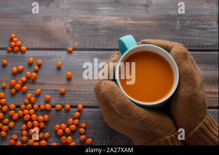 Behandschuhten Händen halten gesund Sanddorn Saft in eine blaue Tasse auf einem Holztisch. Die Beeren sind in der Nähe von Stockfoto