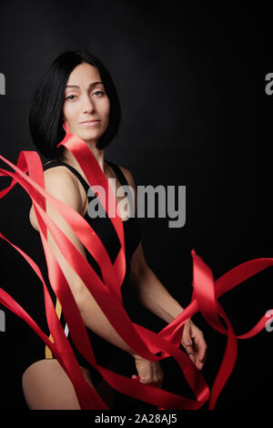 Junge Frau Turnerin der kaukasischen Aussehen mit schwarzem Haar dreht Rot Satin Bänder, gymnastischen Übungen auf schwarzem Hintergrund, selektiver Fokus Stockfoto