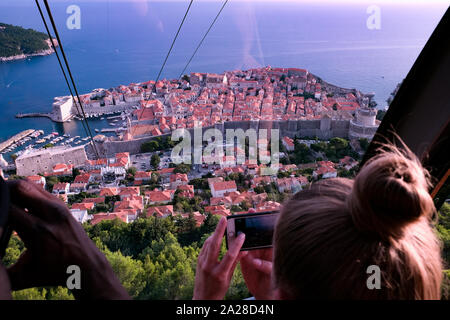 Panoramablick von der Innenseite der Kabine der Seilbahn, die oberhalb der Altstadt von Dubrovnik, die Kabine hoch über der Stadt unten klettert. Ein Tourist nimmt ein Foto Stockfoto