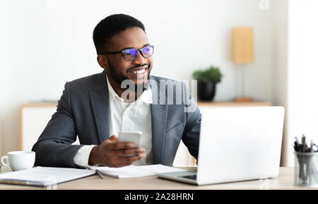 Portrait von lächelnden schwarzer Geschäftsmann am Arbeitsplatz sitzend mit Handy Stockfoto