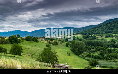 Blick auf die Berge, grüne Wiesen, Bäume, Wälder an den Hängen der Hügel und Bauernhäuser mit einem bewölkten Himmel im Hintergrund. Brens, Frankreich. Stockfoto