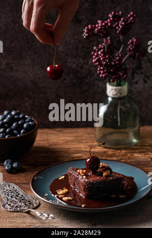 Kaukasische männliche Hand reife Kirsche über hausgemachte vegan Chocolate Brownie, mit Schokoladensauce, auf blauem Teller gekrönt. Holztisch, dunkle b Stockfoto