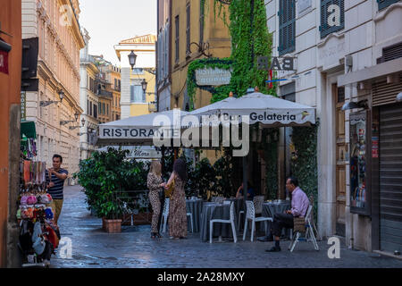 Restaurant im Freien auf einem schmalen, sonnenbeschienene Straße von Rom unter massiven Sonnenschirme Set mit zwei Damen suchen Wie andere Gäste essen. Shop auf der anderen Seite. Stockfoto