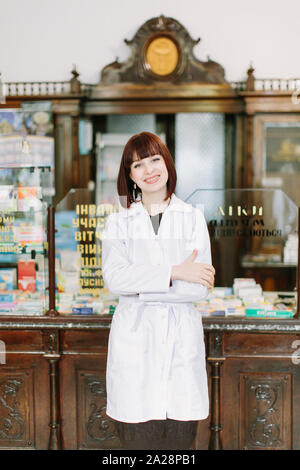 Selbstbewussten jungen Frau Apotheker mit einem reizenden Lächeln stand mit verschränkten Armen in der Apotheke. Stockfoto