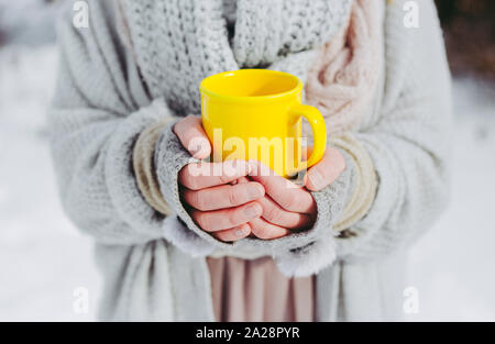 Boho Mädchen, die einen weichen, gewebten Pullover tragen, ist im Winter draußen und hält eine gelbe Tasse warmen Getränks in den Händen. Geringe Schärfentiefe. Stockfoto