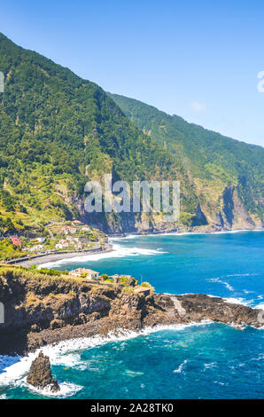 Schönen Klippen an der Küste des Atlantischen Ozeans durch Dorf Seixal, Madeira, Portugal. Steilen grünen Felsen, kleine Stadt und blaues Meer Wasser auf vertikale Fotografie festgehalten. Touristische Destination. Stockfoto