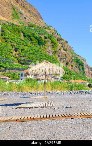 Regenschirm gegen die Sonne auf einem Stein Strand mit Bananenplantagen, auf einem Hügel im Hintergrund auf vertikale Fotografie festgehalten. In Madalena, Mar, Madeira, Portugal. Stockfoto