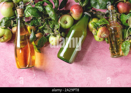 Vielzahl an Apple Getränke. Flaschen Apfelsaft, Essig und Apfelwein mit Garten Äpfel mit Blättern und Zweigen über rosa Textur Hintergrund. Flach, Stockfoto