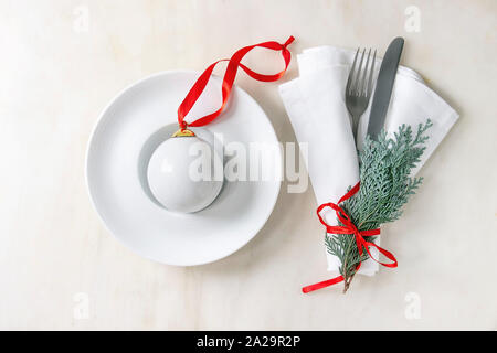 Weißes Porzellan Weihnachtskugel mit roter Schleife in Keramik Teller mit Gabel, Messer, Serviette und thuja Zweig über weißem Marmor Hintergrund. Flach, spac Stockfoto