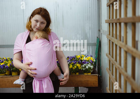 Mutter, die schlafenden Säugling Tochter in einer Schlinge wickeln. Babywearing während Botengänge und Einkaufen in einem Gewächshaus. Stockfoto