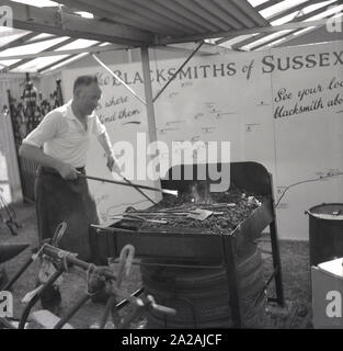 1950er Jahre, ein Schmied, der seine Fertigkeiten in der Eisenschmiede in einem Zelt bei einer landwirtschaftlichen Ausstellung demonstrierte und die Fertigkeiten von Harmern oder Metallarbeitern förderte, Sussex, England, Großbritannien. Auf dem Bild ist eine große Tafel zu sehen, die die Standorte der verschiedenen Schmiede von Sussex zeigt. Stockfoto