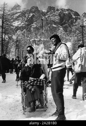 Das Bild zeigt die Besucher der Olympischen Winterspiele 1956 in Cortina d'Ampezzo. Eine Dame in einem Rollstuhl und ihren beiden Gefährtinnen steht im Schnee zwischen den anderen Fans, Sportler, und drücken Sie die Menschen. Stockfoto