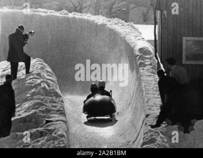 Bei den Olympischen Winterspielen 1956 in Cortina d'Ampezzo 4-Mann und 2-Mann Bob Wettbewerbe ausgetragen wurden. Das Bild zeigt ein Team in einem zwei-Bob während der Fahrt. Ein Fotograf hat sich auf den Rand der Bobbahn geklettert (links). Stockfoto