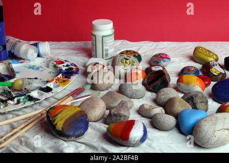 Eine Auswahl bemalter Steine auf einem Basteltisch mit Pinsel für die Felsmalerei als Teil der Rockfun-Phänomene auf rotem Hintergrund Stockfoto