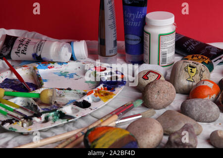 Eine Auswahl von bemalten Felsen auf einer Kunst und Handwerk Tabelle mit Bürsten setup für Rock Malerei als Teil der Rockfun Phänomene auf rotem Hintergrund Stockfoto