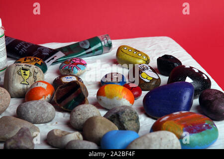 Eine Auswahl bemalter Steine auf einem Basteltisch mit Pinsel für die Felsmalerei als Teil der Rockfun-Phänomene auf rotem Hintergrund Stockfoto
