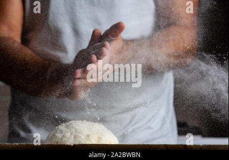 Man knetet Rohteig, Staub aus Mehl in verschiedene Richtungen, männliche Hände auf einem schwarzen Hintergrund fliegt Stockfoto